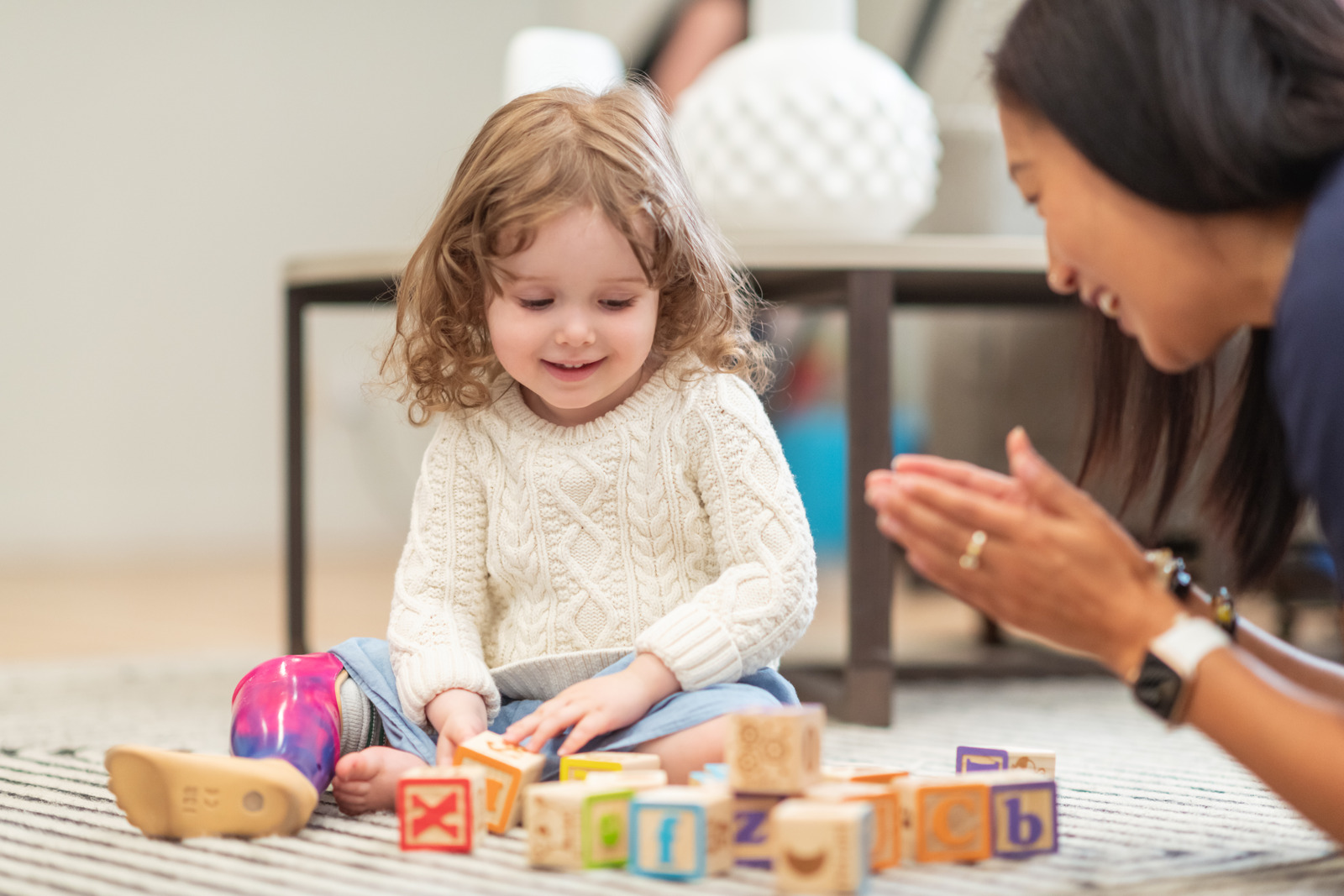 Child with a prosthetic leg plays with wooden blocks on a carpet, while an adult claps and encourages them nearby.
