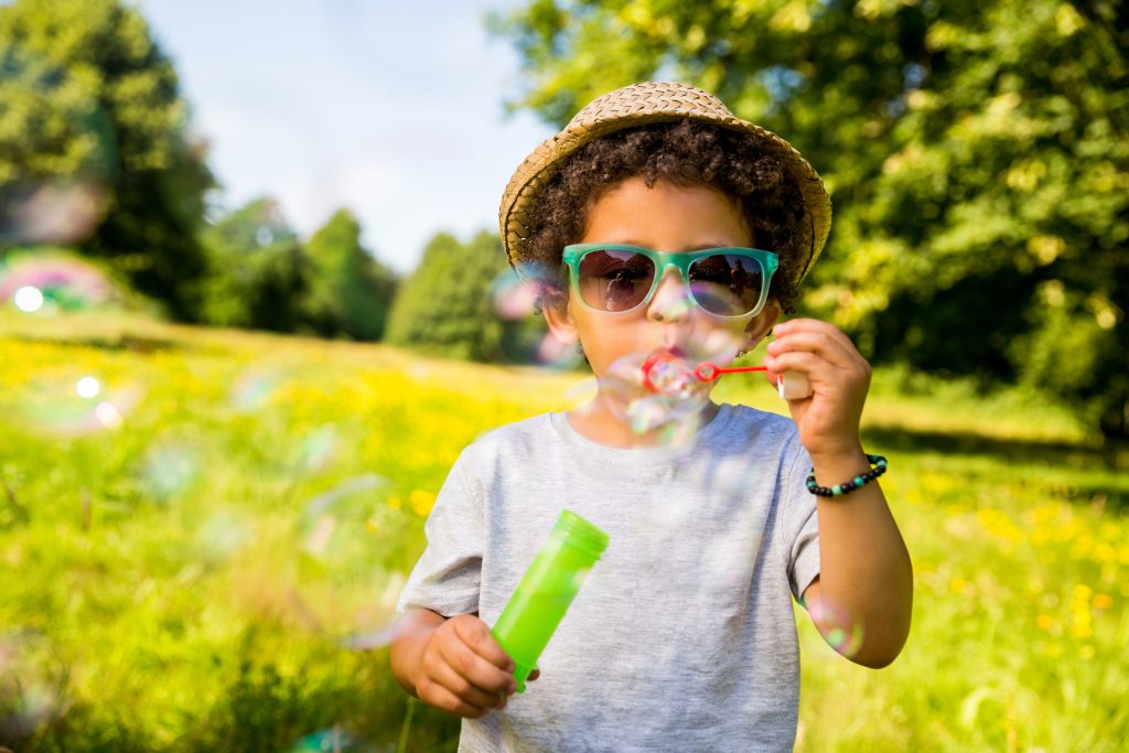 Child wearing a hat and sunglasses blowing bubbles in a sunny, green park with trees in the background.