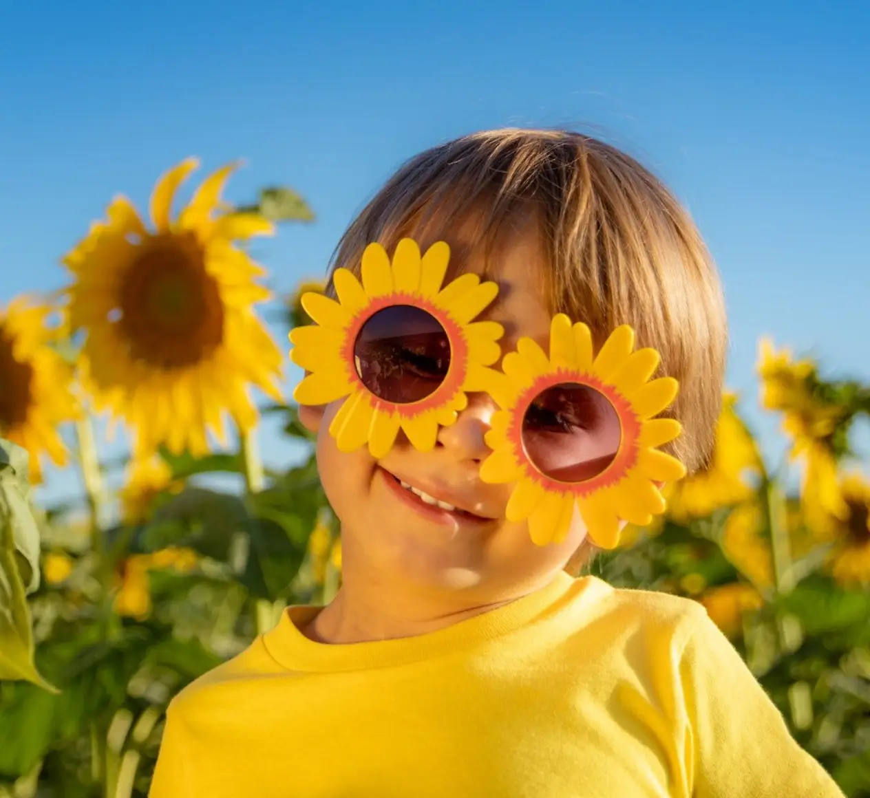 Child wearing sunflower-shaped sunglasses smiles in a sunflower field under a clear blue sky.