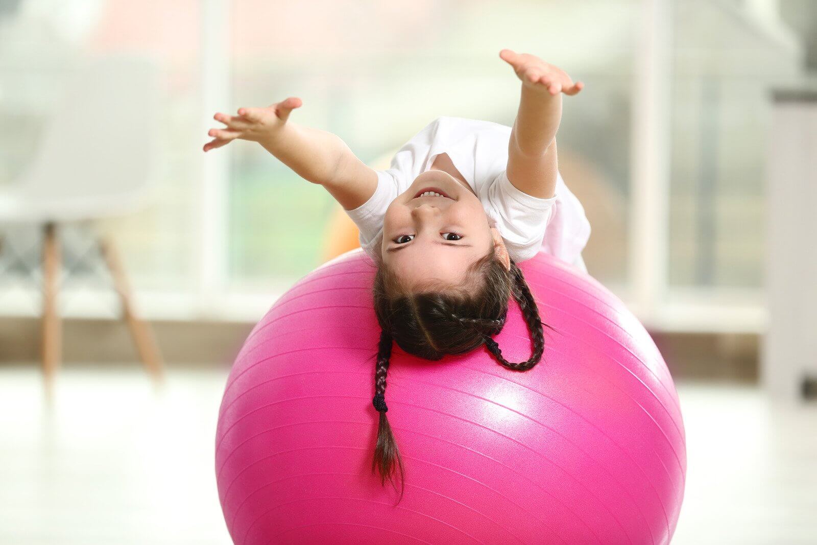 Child with braids lying backwards on a pink exercise ball, smiling and reaching upwards in a bright room.
