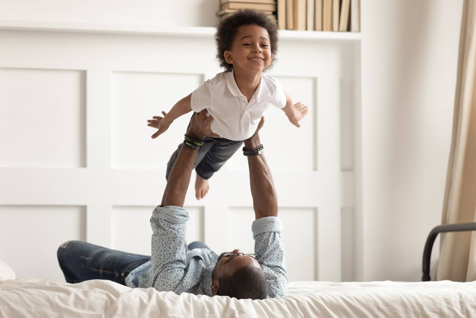 A man lying on a bed lifts a smiling child in the air. The child spreads their arms wide. The setting is a bright room with a white paneled wall and shelves.