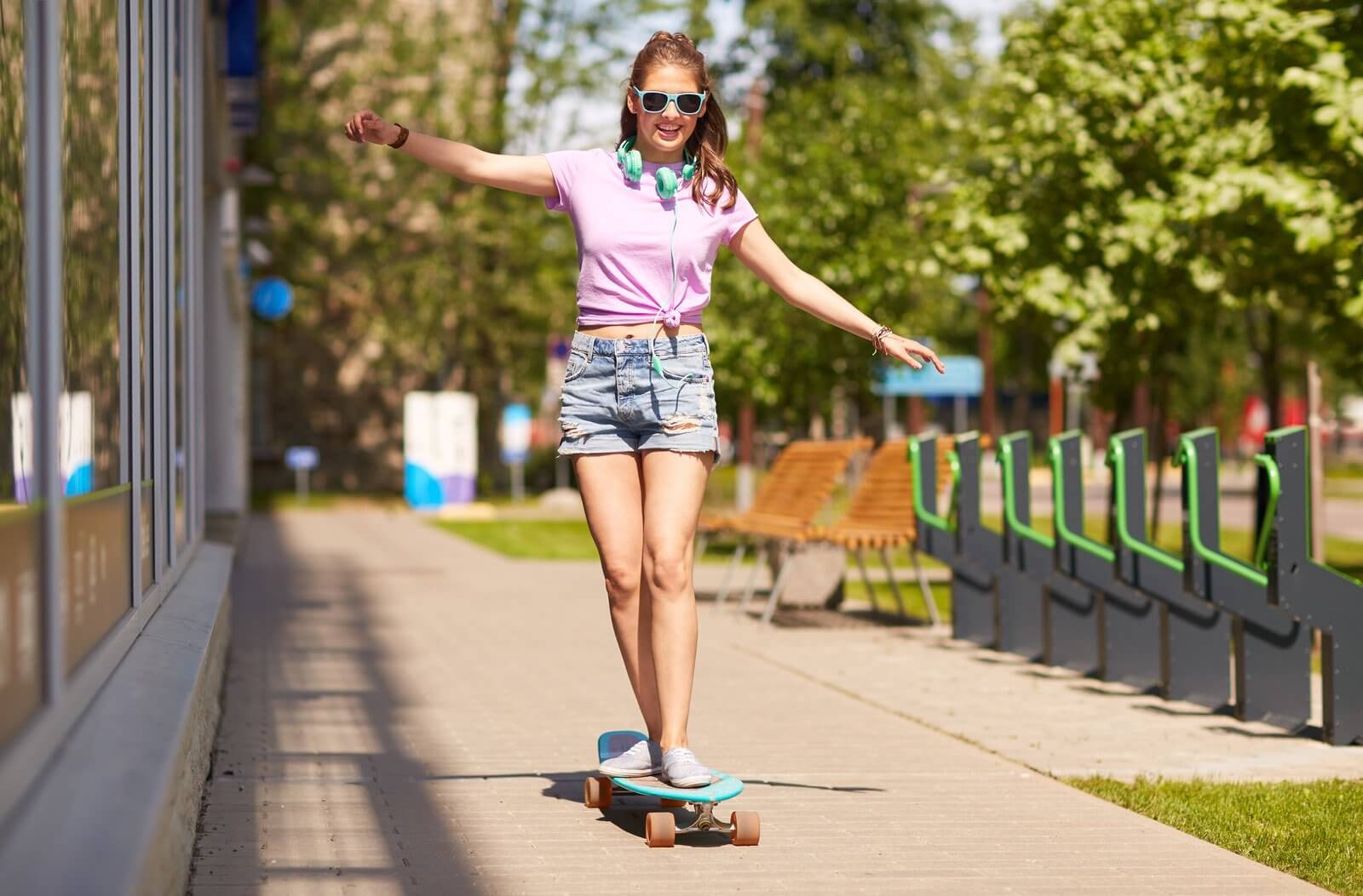 Person wearing sunglasses and headphones skateboards on a sidewalk, balancing with arms outstretched.