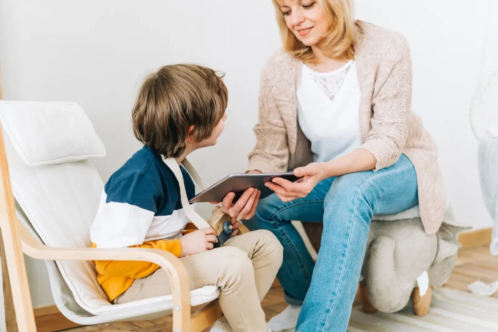 A woman kneels beside a young boy seated in a chair. They are looking at a tablet, and the interaction appears focused and engaged.