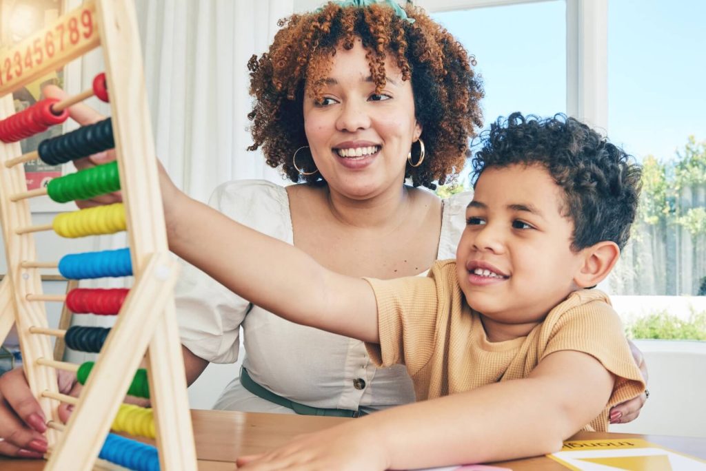 A woman and child use an abacus together, sitting at a table by a window with sunlight streaming in.