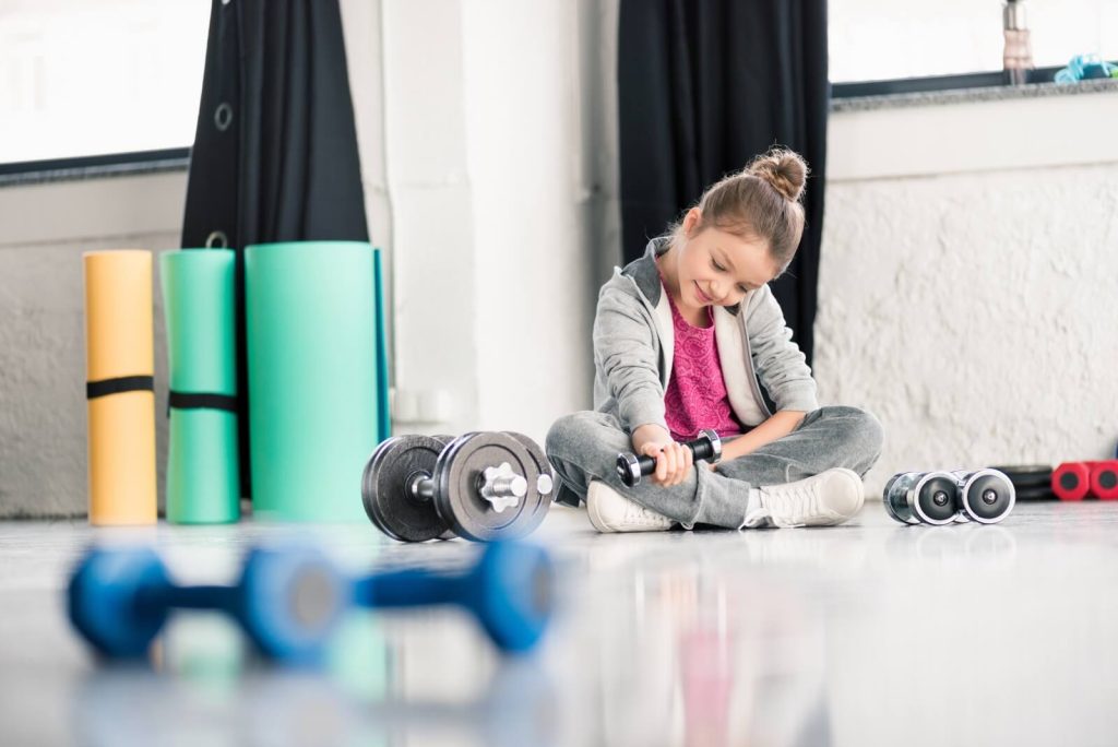 A young girl sits on a gym floor, holding a dumbbell. Several other weights and yoga mats are scattered around her.