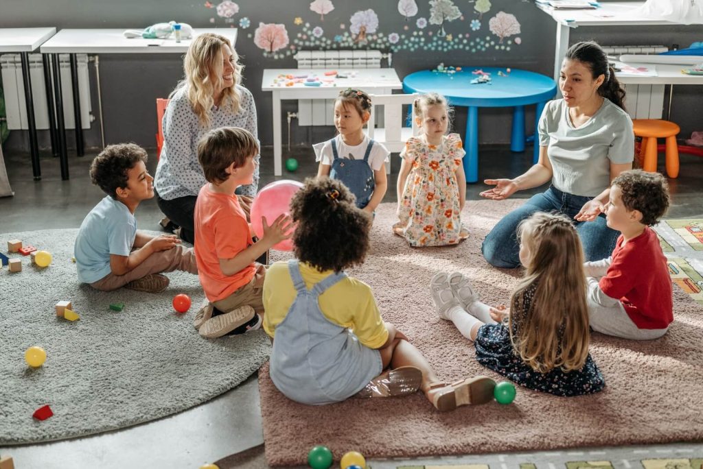 Children and two adults sit in a circle on a carpet in a classroom, surrounded by toys and colorful tables.