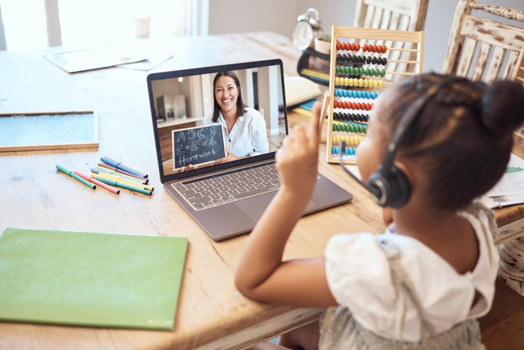 Child wearing headphones participates in a virtual learning session on a laptop. An abacus and colored pencils are on the table.