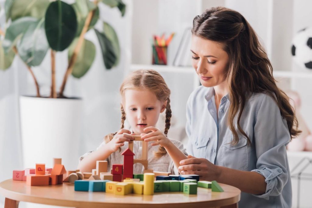 A woman and child with braided hair are sitting at a table, playing with colorful wooden blocks. A potted plant and a shelf with items are in the background.