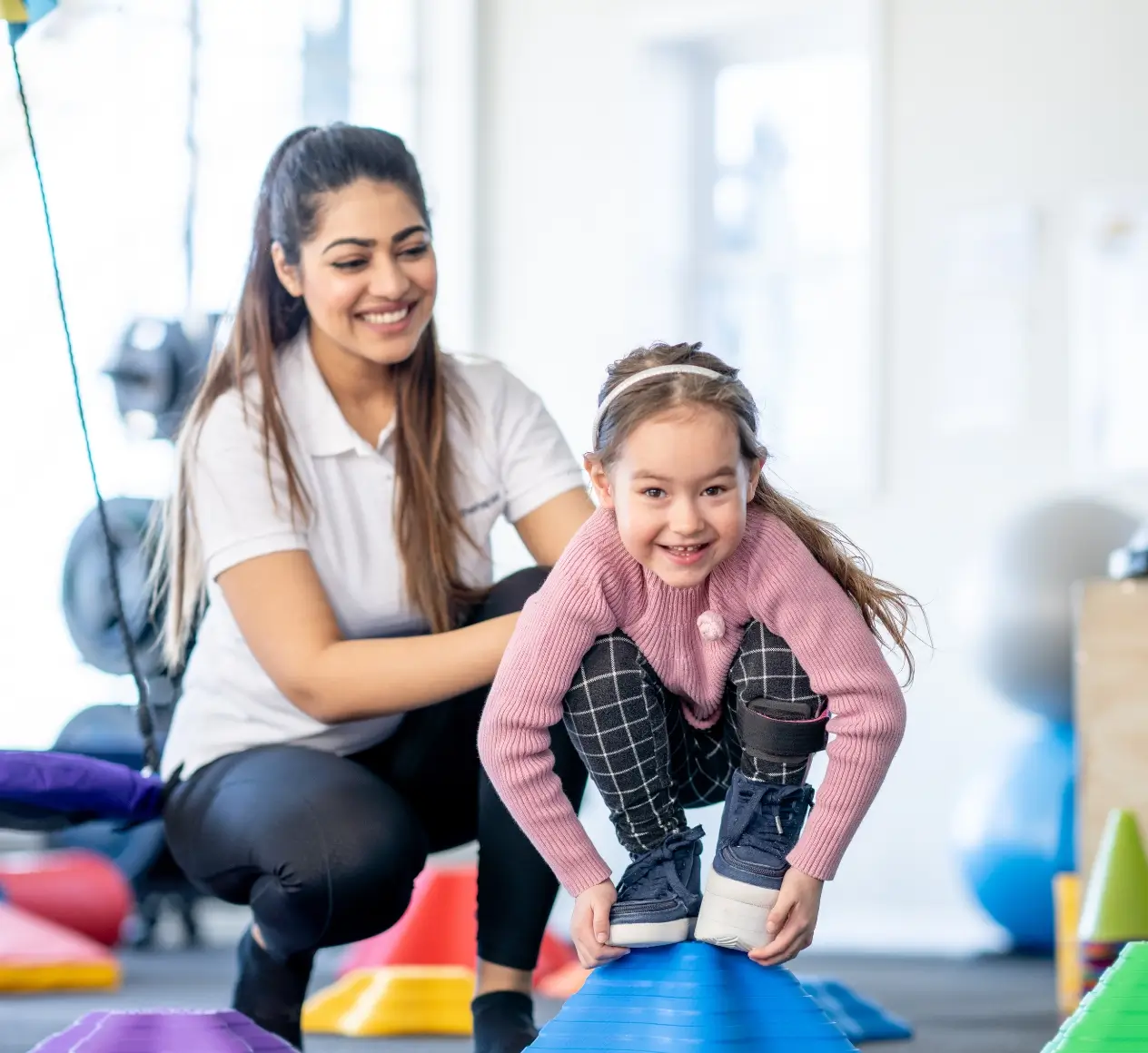 A woman assists a young girl balancing on a blue, cone-shaped object in a gym, surrounded by colorful exercise equipment.