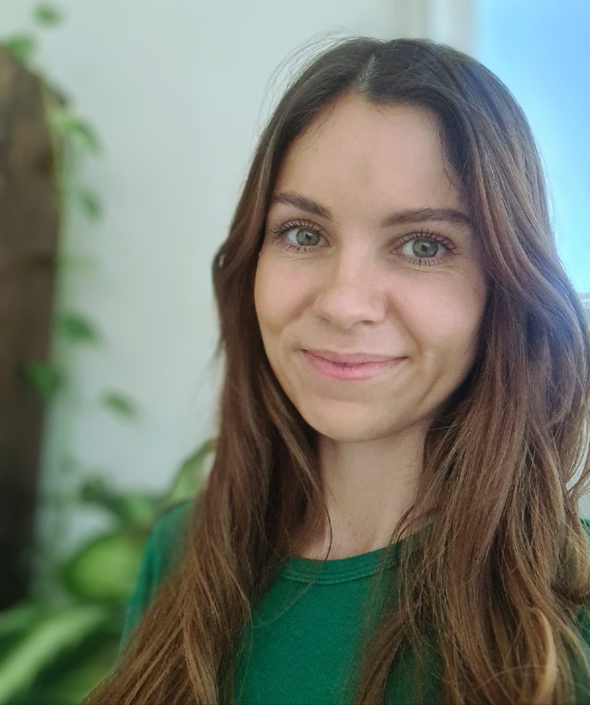 A person with long brown hair and a green shirt smiles at the camera. There is a blurred background with greenery.